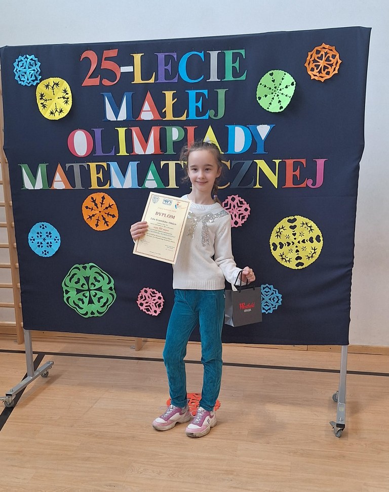 Julia Krzemińska-Nikścin na tle baneru z napisem: 25-lecie Małej Olimpiady Matematycznej, prezentuje zdobyte trofea- dyplom i nagrodę.
